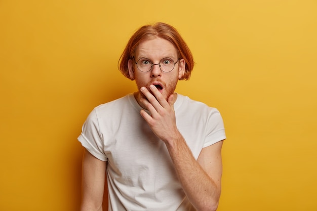 Hombre pelirrojo aturdido reacciona sobre los descuentos de temporada, mira con estupor, se tapa la boca, usa gafas y camiseta blanca, aislado en la pared amarilla, se olvidó de algo. Omg expresión