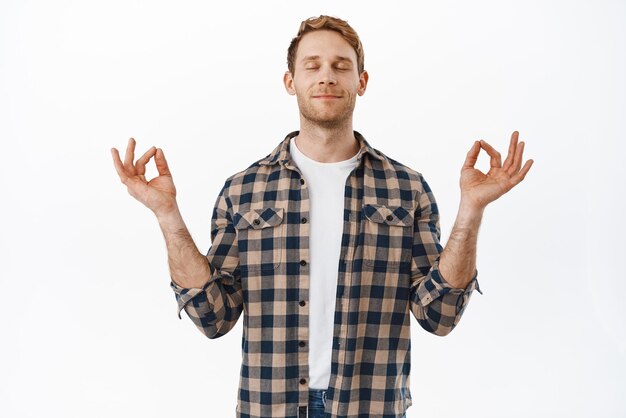 El hombre pelirrojo cierra los ojos y medita con mudras zen sonriendo y respirando meditación pacífica relajada o yoga tratando de calmarse de pie sobre fondo blanco