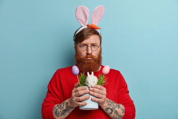 Foto gratuita el hombre pelirrojo descontento sonríe y se ve infeliz, ha estropeado el día, posa con un pequeño conejito de pascua, huevos coloridos decorados, usa anteojos redondos, orejas de conejo, posa con expresión sombría