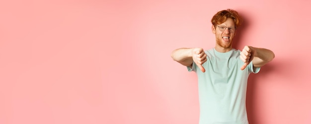 Hombre pelirrojo disgustado con camiseta y gafas mostrando los pulgares hacia abajo y la lengua haciendo muecas de aversión