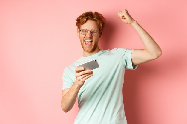 Hombre pelirrojo feliz ganando en videojuegos para móviles, levantando la mano y gritando sí con alegría, celebrando la victoria, mirando el teléfono inteligente, de pie sobre fondo rosa.