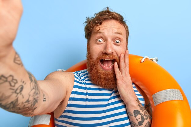 Hombre pelirrojo positivo divertido se toma una foto, se para con un salvavidas inflado en el interior, vestido con chaleco de marinero, tiene una barba espesa, disfruta de unas vacaciones increíbles en la playa