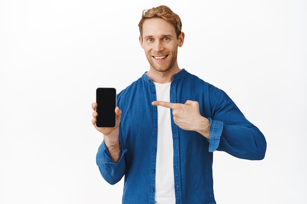 Hombre pelirrojo sonriente confiado apuntando a la pantalla del teléfono y mirando al frente, recomendando una buena aplicación en el teléfono inteligente, muestra la oferta comercial, pared blanca