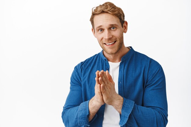 El hombre pelirrojo sonriente hace un gesto de oración y se ve agradecido, aprecia la ayuda, agradece, expresa gratitud, de pie sobre una pared blanca