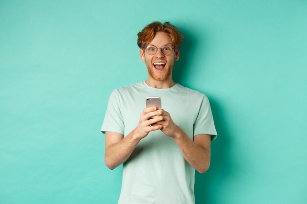 Hombre pelirrojo sorprendido con gafas mirando asombrado a la cámara después de leer la oferta promocional en el teléfono inteligente, de pie contra el fondo turquesa.