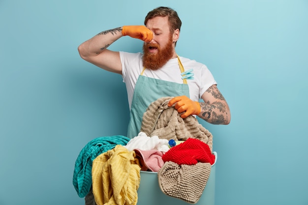 Foto gratuita el hombre pelirrojo se tapa la nariz, siente mal olor, olor desagradable de la ropa sucia, va a lavarse con polvo líquido, usa guantes de goma y delantal, está ocupado haciendo las tareas del hogar durante el fin de semana. ¡qué hedor!