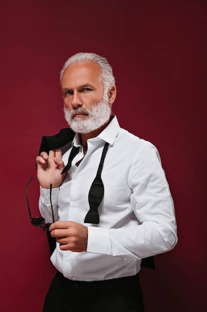 Hombre de pelo blanco en camisa sostiene anteojos Chico adulto moderno con barba en traje clásico mirando a la cámara sobre fondo aislado