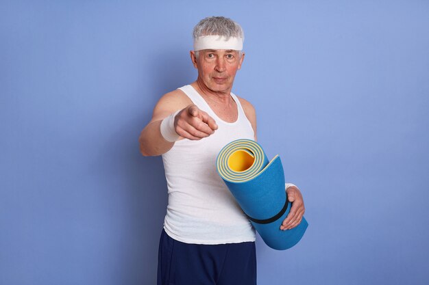 Hombre de pelo blanco senior deportivo sosteniendo una estera de yoga, mirando y apuntando al frente