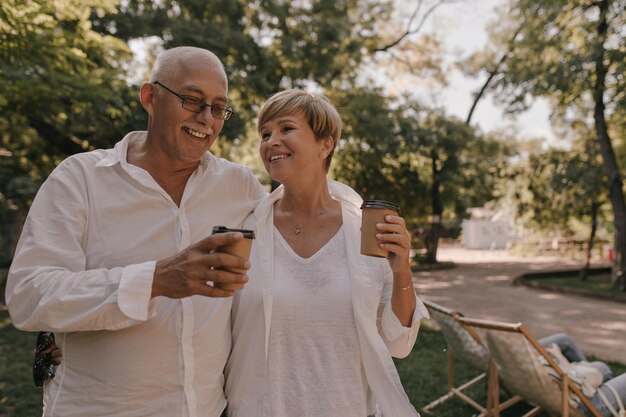 Hombre de pelo gris con anteojos en camisa blanca sonriendo y posando con gorra de té y mujer elegante en traje ligero al aire libre