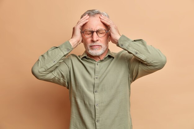 Hombre de pelo gris disgustado sufre dolor de cabeza mantiene las manos en la cabeza para revelar el dolor necesita analgésicos tiene migraña después de que una fiesta ruidosa usa camisa formal aislada sobre una pared marrón