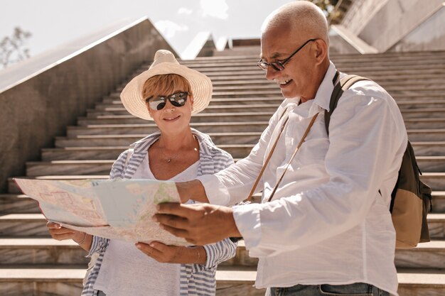 Hombre de pelo gris con gafas y camisa ligera con mochila mirando el mapa con una mujer moderna con sombrero y ropa de rayas azules