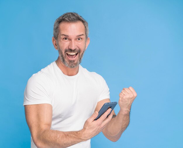 Hombre de pelo gris de mediana edad con teléfono inteligente en la mano feliz sonriendo a la cámara con camiseta blanca aislada sobre fondo azul
