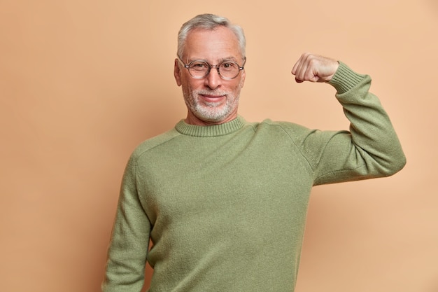 Hombre de pelo gris satisfecho y confiado levanta el brazo y muestra el músculo demuestra resultados después de un entrenamiento regular en el gimnasio usa anteojos y un puente aislado sobre una pared marrón estando orgulloso de sí mismo