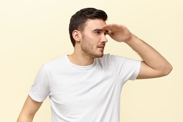 Hombre de pelo oscuro joven lindo divertido en camiseta casual sosteniendo la palma en la frente y entrecerrar los ojos