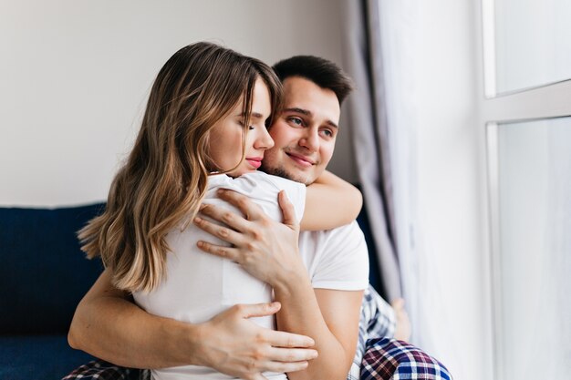 Hombre de pelo oscuro romántico abrazando a su esposa. Filmación en interiores de feliz chica rubia relajándose en un acogedor sofá.