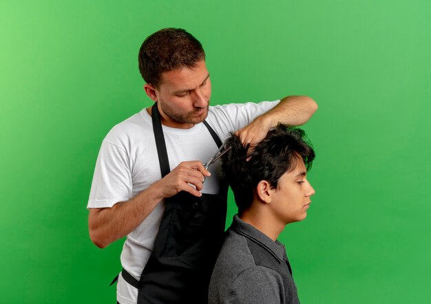 Hombre de peluquero en delantal cortando el cabello con tijeras de cliente satisfecho de pie sobre la pared verde 3