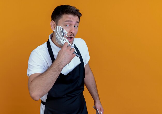 Hombre de peluquero en delantal con espuma de afeitar en la barba afeitándose con navaja de pie sobre fondo naranja