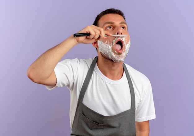 Hombre de peluquero en delantal con espuma de afeitar en la cara afeitándose con navaja de pie sobre la pared púrpura