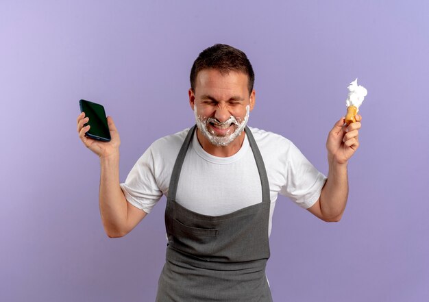 Hombre de peluquero en delantal con espuma de afeitar en su rostro sosteniendo smartphone y brocha de afeitar mirando emocionado y feliz de pie sobre la pared púrpura