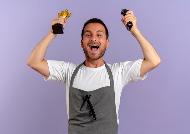 Hombre de peluquero en delantal con máquina de corte de pelo y trofeo feliz y emocionado regocijándose de su éxito de pie sobre la pared púrpura
