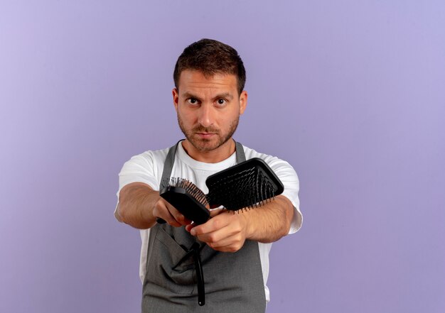 Hombre de peluquero en delantal sosteniendo cepillos para el cabello mirando hacia el frente con rostro serio de pie sobre la pared púrpura