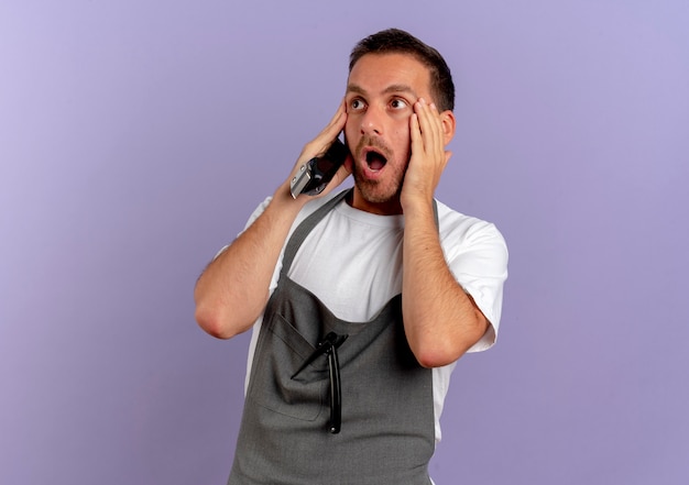 Hombre de peluquero en delantal sosteniendo la máquina de corte de cabello mirando a un lado sorprendido con la boca abierta de pie sobre la pared púrpura