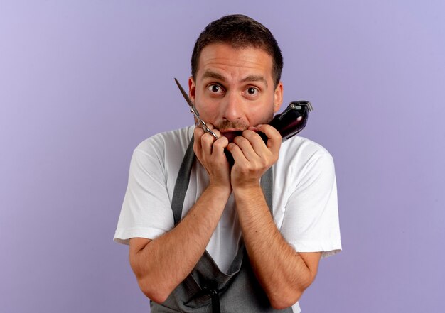 Hombre de peluquero en delantal sosteniendo la máquina de corte de pelo y tijeras estresado y nervioso morderse las uñas de pie sobre la pared púrpura