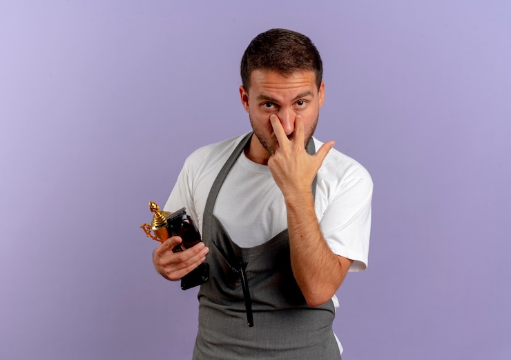 Foto gratuito hombre de peluquero en delantal sosteniendo la máquina de corte de pelo y trofeo apuntando con los dedos a los ojos de pie sobre la pared púrpura
