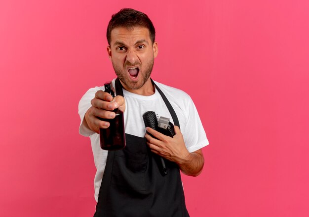 Hombre de peluquero enojado en delantal sosteniendo cepillo de pelo y recortadora apuntando hacia el frente con spray de pie sobre pared rosa