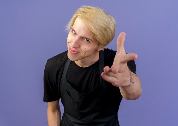 Foto gratuita hombre de peluquero profesional en delantal apuntando con el brazo a usted sonriendo de pie sobre la pared azul