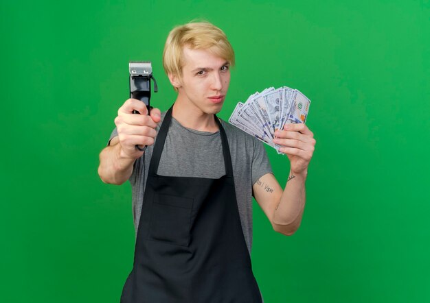 Hombre de peluquero profesional en delantal con efectivo mostrando recortador mirando confiado