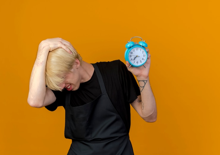 Foto gratuito hombre de peluquero profesional en delantal con reloj despertador confundido y muy ansioso de pie sobre la pared naranja