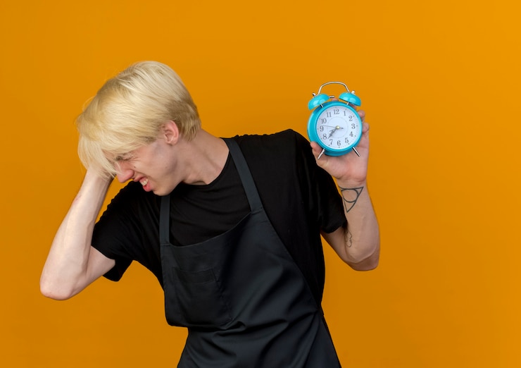 Foto gratuito hombre de peluquero profesional en delantal con reloj despertador confundido y muy ansioso