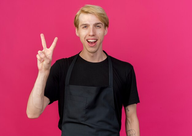 Hombre de peluquero profesional en delantal sonriendo con cara feliz mostrando v-sign