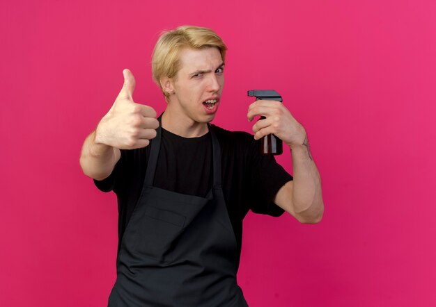 Hombre de peluquero profesional en delantal sosteniendo spray sonriendo confiado mostrando los pulgares para arriba