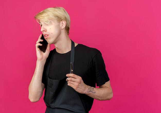 Hombre de peluquero profesional en delantal con tijeras hablando por teléfono móvil confundido