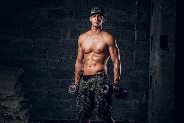 Un hombre pensativo sin camisa con gorra está haciendo ejercicios con pesas en un estudio fotográfico oscuro.