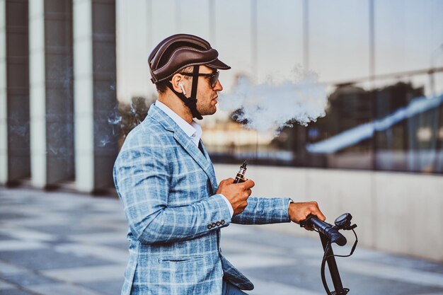 Un hombre pensativo con casco protector y gafas de sol está fumando vape mientras sostiene su scooter eléctrico.