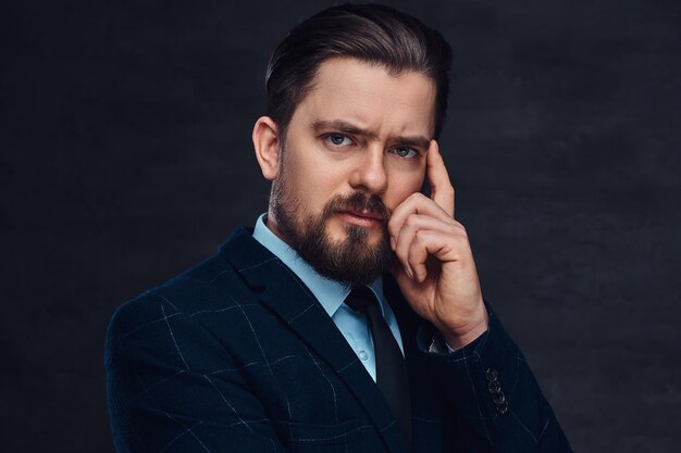 Hombre pensativo de mediana edad con barba y peinado vestido con un elegante traje azul sobre un fondo oscuro texturizado en el estudio.