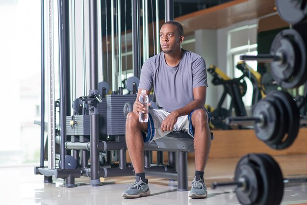 Hombre pensativo sentado en el gimnasio, sosteniendo la botella de agua