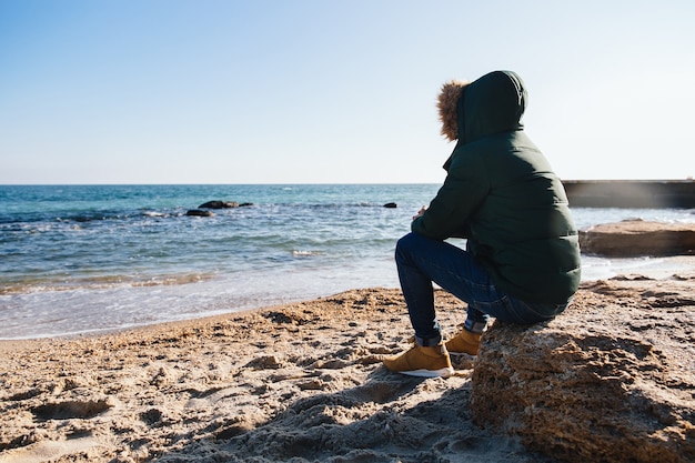 Hombre pensativo sentado en la piedra, mirando el mar. Vestido con una cálida chaqueta con capucha.