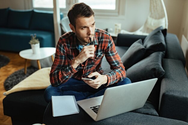 Hombre pensativo usando teléfono móvil y computadora portátil mientras trabaja en casa