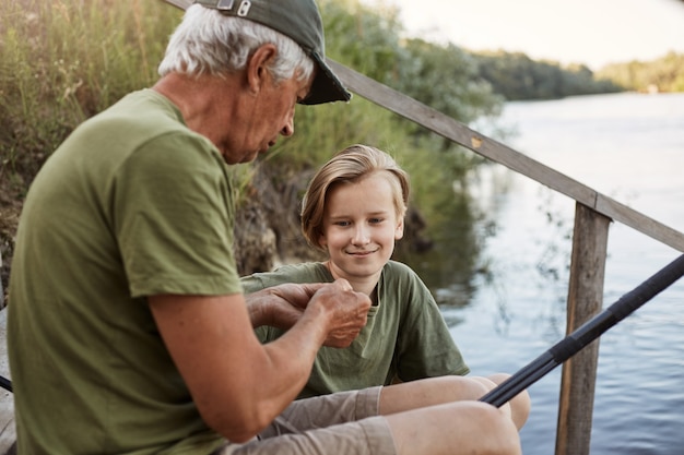 Hombre pescador enseñando a su nieto a anclar el cebo, conocido como método para pescar, un joven rubio sonriente mira a un hombre mayor con una sonrisa y una mirada concentrada, sentarse en las escaleras de madera al agua.