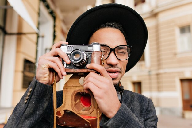 Hombre con piel de color marrón claro tomando fotografías con cámara. Retrato de primer plano al aire libre del fotógrafo masculino negro lleva sombrero en un día frío.