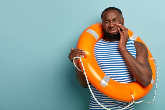 El hombre de piel oscura sin afeitar molesto tiene una expresión facial triste, cierra los ojos, lleva un salvavidas para nadar con seguridad
