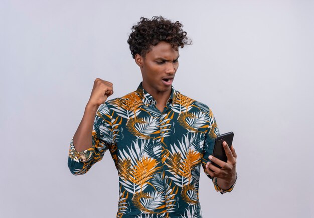 Hombre de piel oscura agresivo y enojado con cabello rizado en hojas camisa impresa perdiendo el juego en el teléfono móvil con los puños apretados