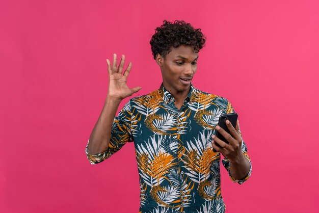 Hombre de piel oscura, guapo y alegre, con cabello rizado en hojas, camisa estampada, hablando con un amigo durante la videollamada y agitando la mano en la cámara frontal del teléfono móvil sobre un fondo rosa