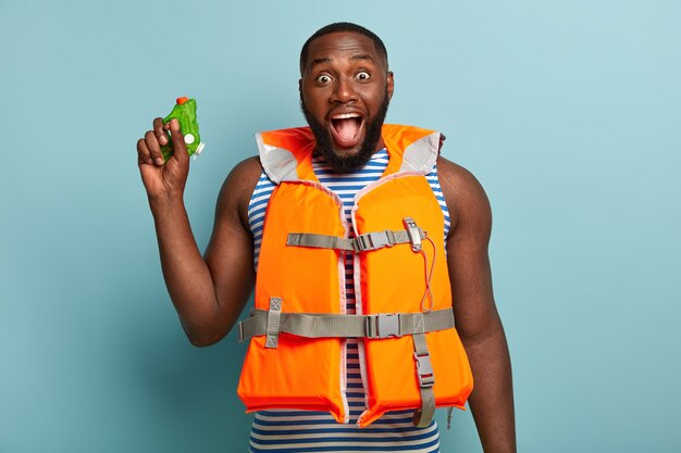 Hombre de piel oscura lleno de alegría tiene agua peleando con amigos en la playa, exclama felizmente, usa chaleco salvavidas naranja