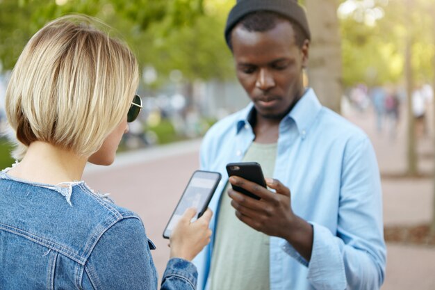 Hombre de piel oscura de moda con sombrero negro y camisa de pie en la calle con teléfono móvil y su amigo rubio, usando internet, intercambiando archivos o fotos. Mejores amigos de raza mixta reunidos en la calle