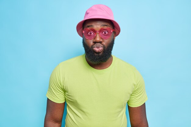 El hombre de piel oscura de moda viste una camiseta verde panamá rosa y gafas de sol en forma de corazón mantiene los labios redondeados tiene una expresión romántica vestida con ropa de verano